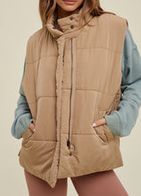 Load image into Gallery viewer, model wearing the long way home puffer vest in the color mocha. model has the vest paired with a long sleeve top in the color sea form and a pair of terracotta leggings.