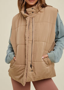 model wearing the long way home puffer vest in the color mocha. model has the vest paired with a long sleeve top in the color sea form and a pair of terracotta leggings.