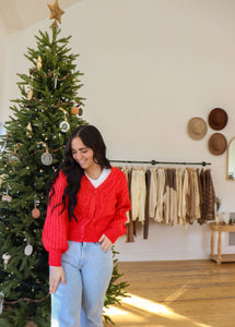 model wearing the where the heart is sweater in the color red. model has the sweater paired with a pair of light wash denim and is posing in front of a Christmas tree.