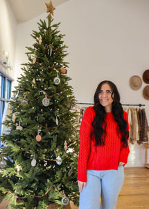 model wearing the my way back home sweater in the color red. model has the sweater paired with a pair of light wash denim and is posing in front of a Christmas tree.