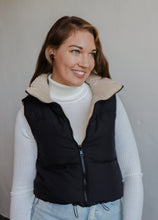 Load image into Gallery viewer, model wearing the vest of both worlds puffer in the color black/taupe. model has the vest paired with the can&#39;t get enough top in the color ivory and a pair of light wash denim.