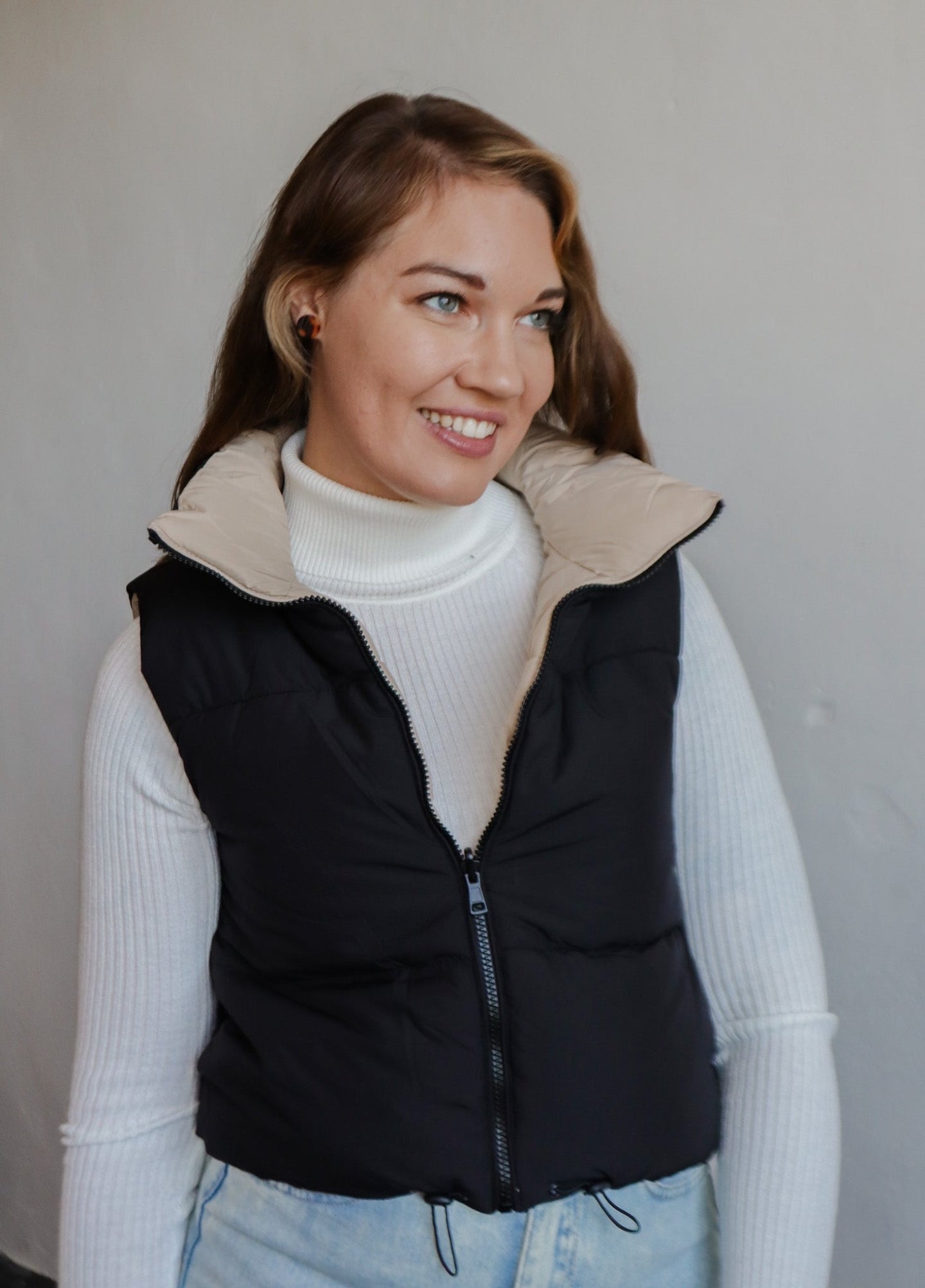model wearing the vest of both worlds puffer in the color black/taupe. model has the vest paired with the can't get enough top in the color ivory and a pair of light wash denim.