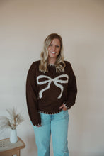 Load image into Gallery viewer, front view of model wearing the ribbons + bows sweater in the color chocolate. model has the sweater paired with the charleston denim.