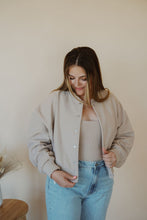 Load image into Gallery viewer, front view of model wearing the winding roads bomber jacket in the color light taupe. model has the jacket paired with the used to know bodysuit in the color biscuit and the charleston denim.