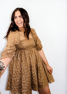 angled view of smiling model wearing the familiar glow dress with one hand pinching the side of the dress and the other holding a bunch of flowers that is mostly out of the camera's view