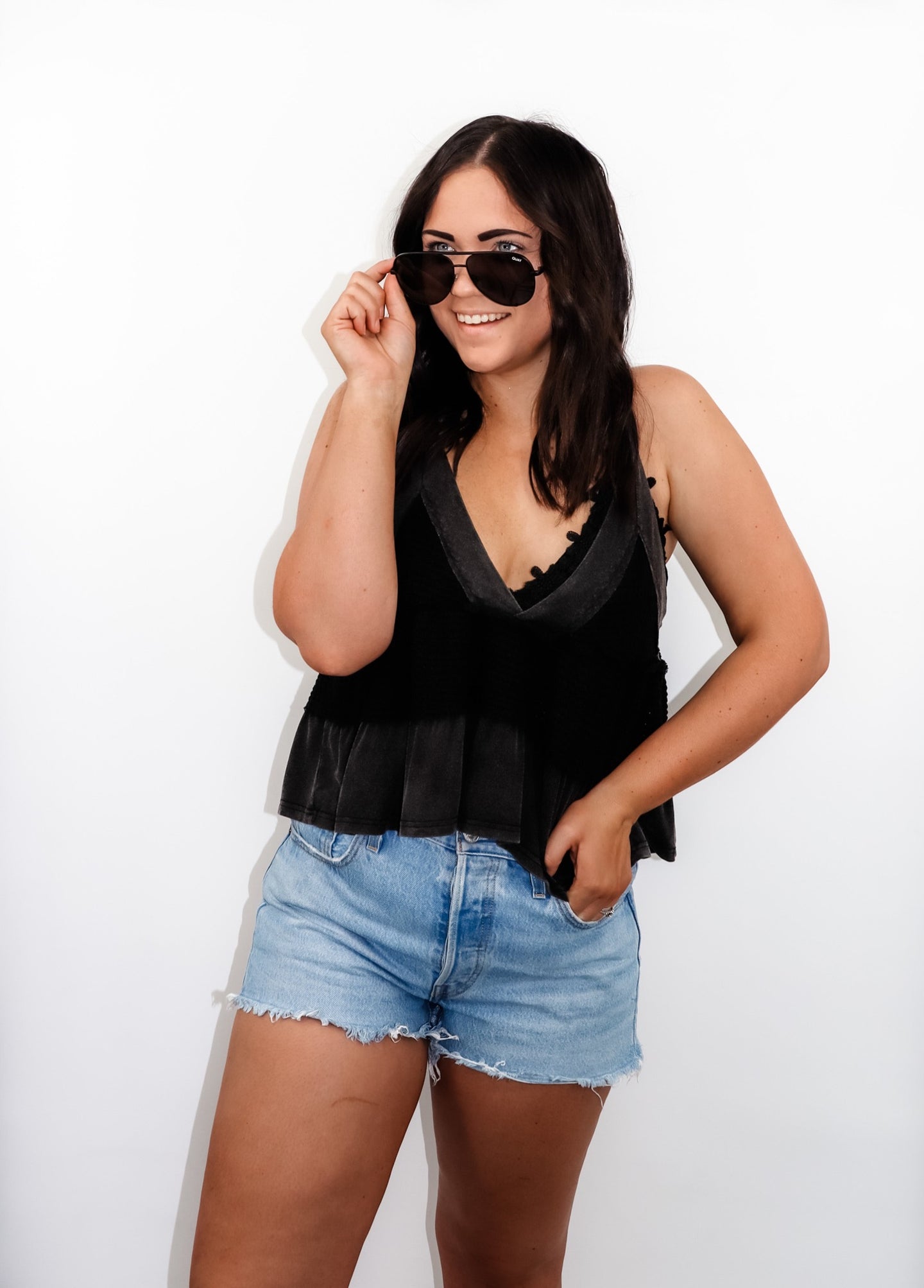 model wearing the moving on top in the color black paired with light denim shorts; model is wearing a pair of sunglasses and has one hand pinching the frames