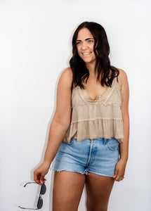 model wearing the moving on top in the color taupe paired with light denim shorts; model is holding a pair of sunglasses in one hand