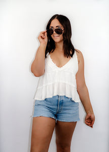 model wearing the moving on top in the color white paired with light denim shorts; model is wearing a pair of sunglasses and has one hand pinching the frames