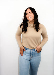 model wearing the by your side top in the color taupe paired with a pair of lightwash denim. model is standing with one hand in her pocket with her thumb through a belt loop and the other hand resting on her thigh. model is smiling and looking to the side.