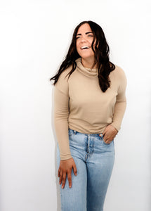 model wearing the by your side top in the color taupe paired with a pair of lightwash denim. model is standing with one hand in her pocket with her thumb through a belt loop and the other hand resting on her thigh. model appears to be laughing and looking to the side.