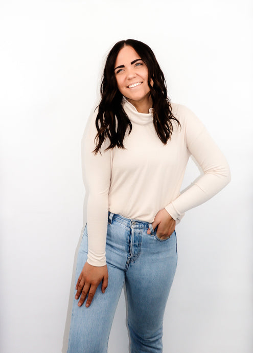 model wearing the by your side top in the color cream paired with a pair of lightwash denim. model is standing with one hand in her pocket with her thumb through a belt loop and the other hand resting on her thigh. model is smiling and looking to the side.