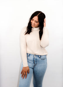 model wearing the by your side top in the color cream paired with a pair of lightwash denim. model is standing with one hand touching her hair and the other resting on her thigh. model is smiling and looking downward towards the ground.