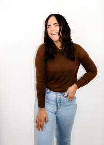 model wearing the by your side top in the color espresso paired with a pair of lightwash denim. model is standing with one hand in her pocket with her thumb through a belt loop and the other hand resting on her thigh. model appears to be laughing and is looking out past the camera.