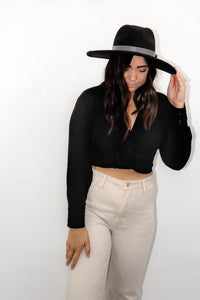 model wearing the all this time top paired with a pair of cream colored denim jeans and a black rancher style hat. model is smiling, looking off toward the side, and resting one hand on her hat and the other on her thigh.