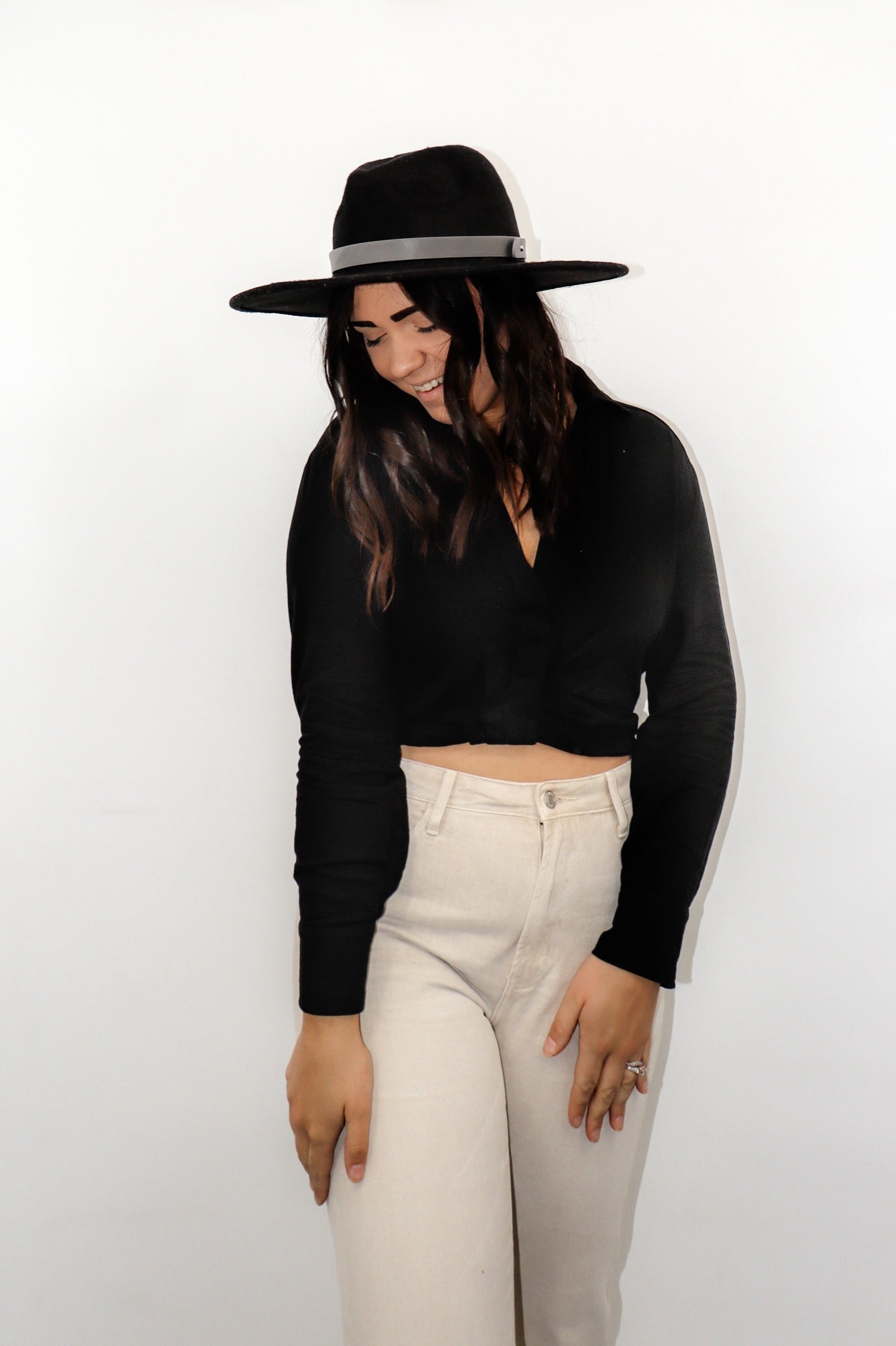 model wearing the all this time top paired with a pair of cream colored denim jeans and a black rancher style hat. model is smiling, looking downward and off toward the side, and resting her hands on her thighs.