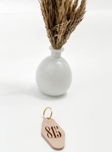 photo of leather motel style keychain with 815 markings on the face of the keychain and gold hardware; keychain is laying in front of a white vase with faux grass stems