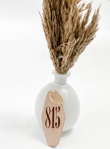 photo of leather motel style keychain with 815 markings on the face of the keychain and gold hardware; keychain is leaning against a white vase with faux grass stems