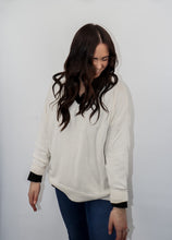 Load image into Gallery viewer, model wearing the state of grace sweater in the color cream. model has the sweater paired with the play by my rules top in the color black and a pair of high-waisted dark-wash denim.